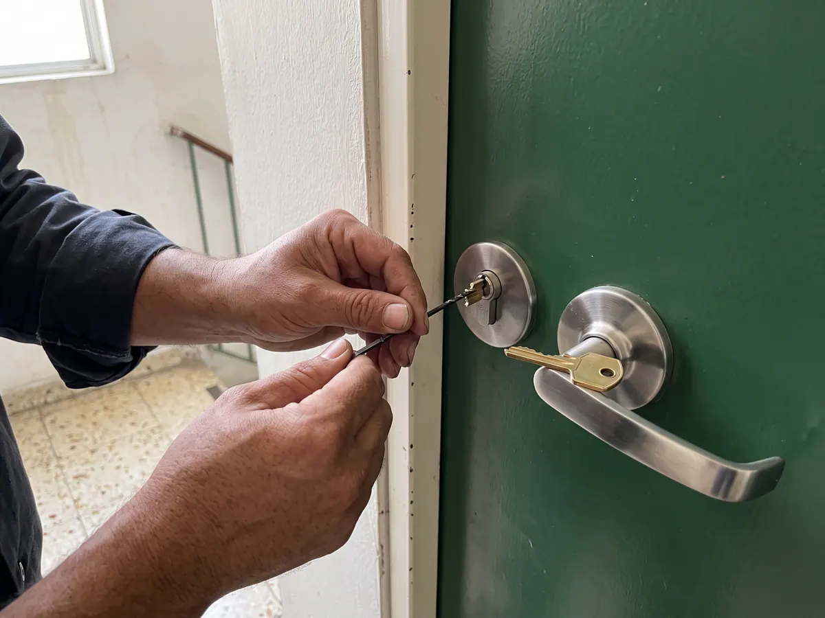 Manos de un cerrajero manipulando un cilindro de puerta con ganzúa y llave de tensión, con el maletín de herramientas abierto en el suelo
